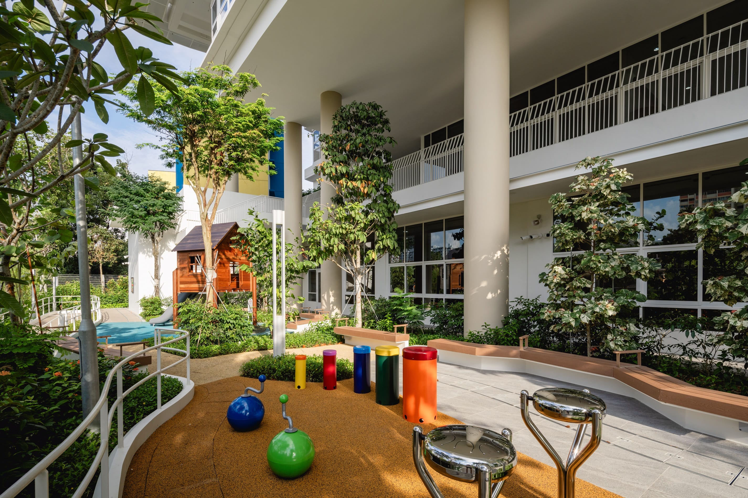 Outdoor playground with playhouse, colorful drums, musical instruments, and trees near a modern building.